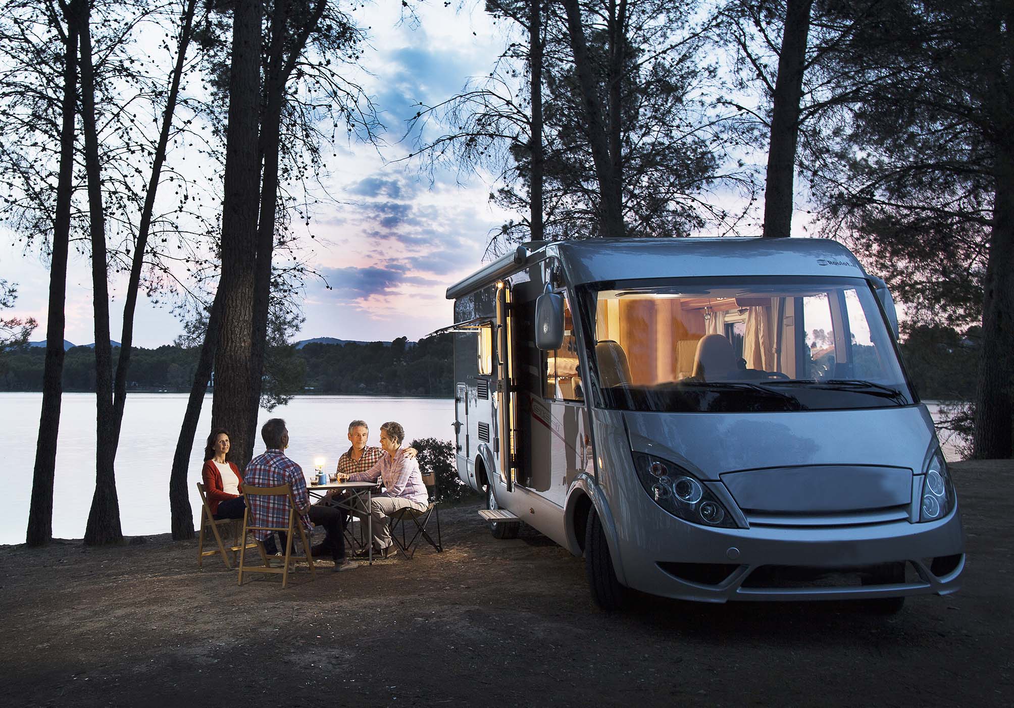 An RV parked next to a lake, with a group of people sitting around a table.