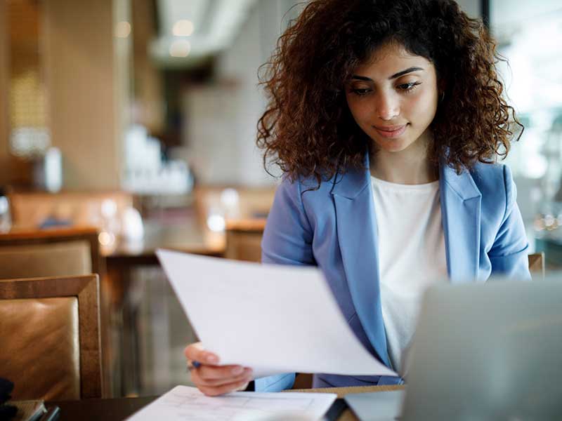 Woman wearing blue sweater sitting at a restaurant while looking at piece of paper. 