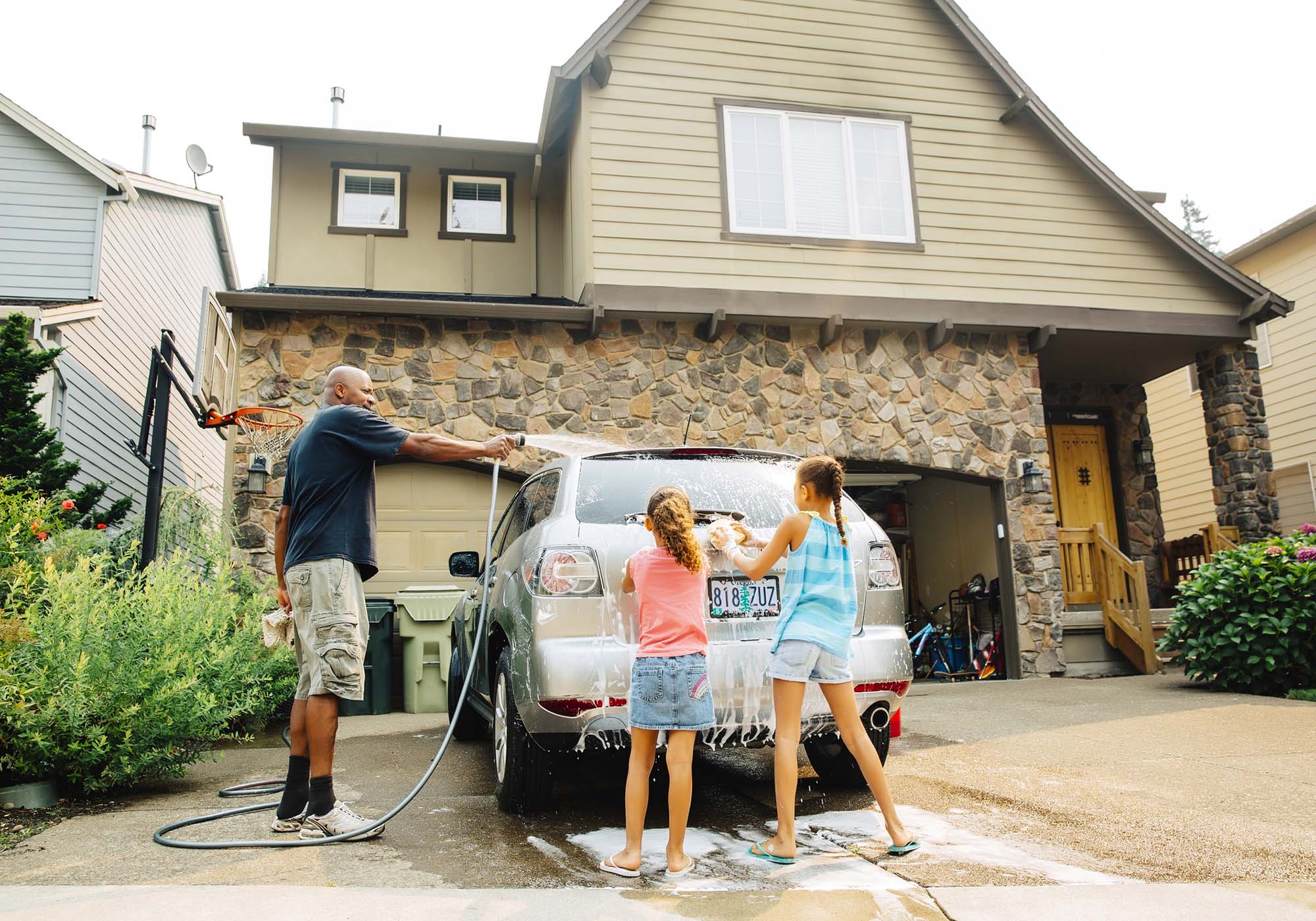 Dad with two daughters washing their silver SUV outside of their house. 