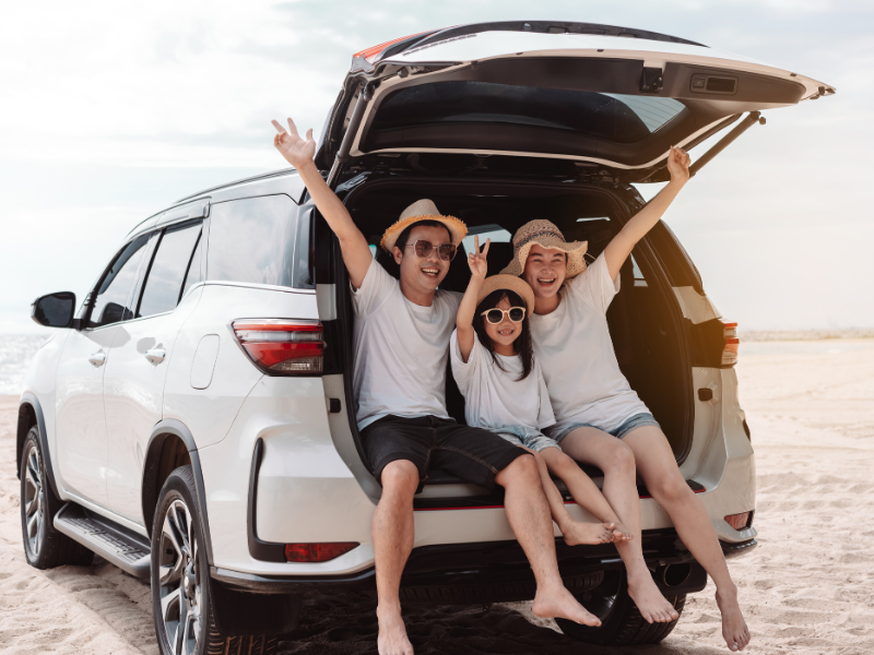 Happy family sitting in the open trunk of an SUV parked at the beach, smiling and enjoying the sunny day together.