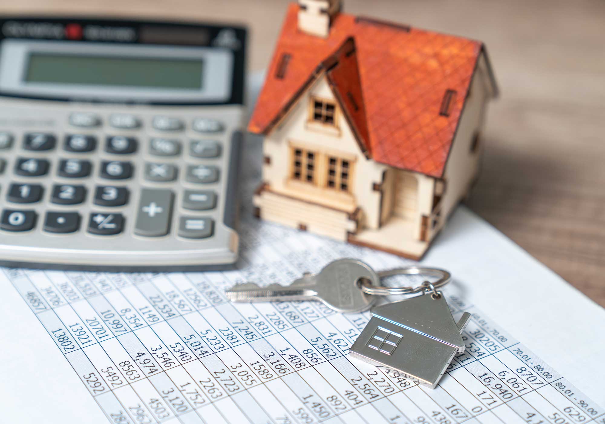 A paper with numbers and a calculator, small model house and key on top of it.