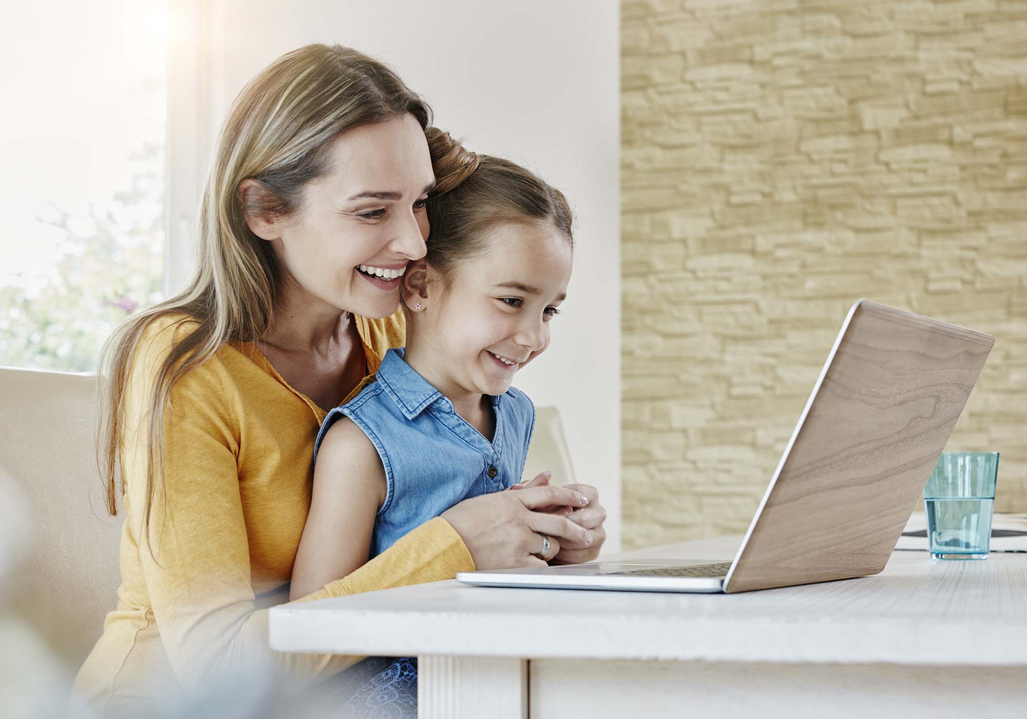 A daughter sitting on her mother's lap, while mother is looking at laptop. 