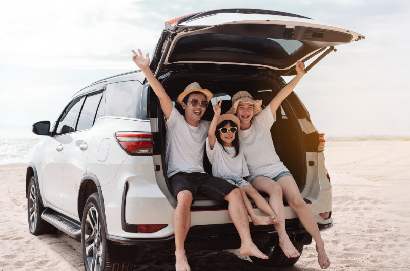 Happy family sitting in the open trunk of an SUV parked at the beach, smiling and enjoying the sunny day together.