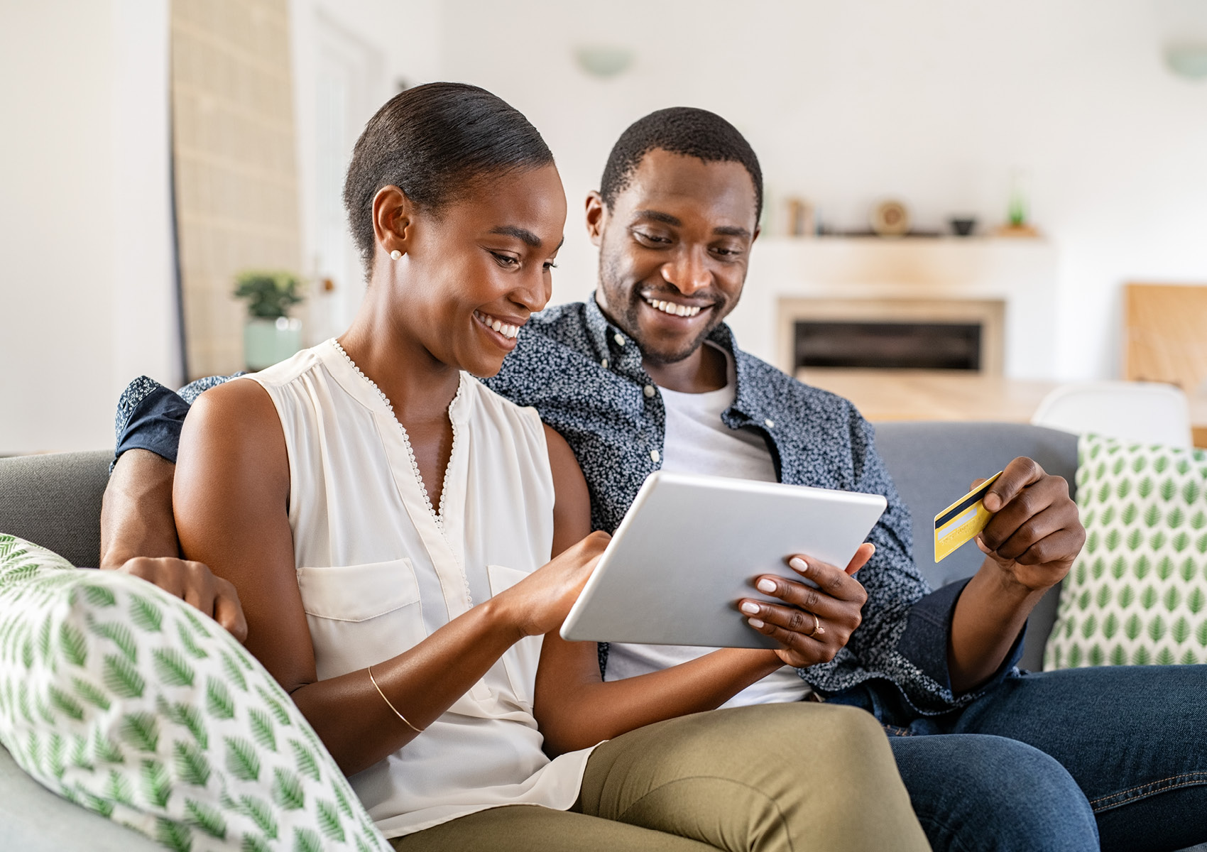 A happy couple sitting on their sofa, the woman is holding an iPad and the man is holding his The Police Credit Union debit card.